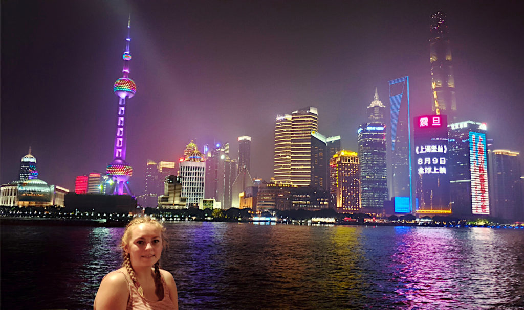 Zoe stood on The Bund in Shanghai with high rise buildings illuminated at night on the other side of the river