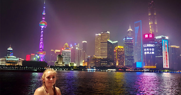 Zoe stood on The Bund in Shanghai with high rise buildings illuminated at night on the other side of the river