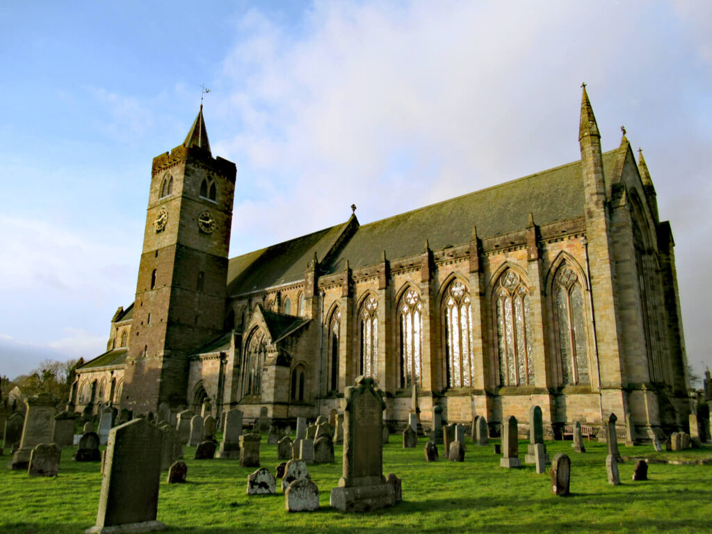 Showing Dunblane Cathedral from the side. Tombstones are shown in the grounds of the church in the foreground of the picture. The cathedral dominates the picture with stunning brickwork and stained glass windows. A clock tower is visible on the left side of the church.