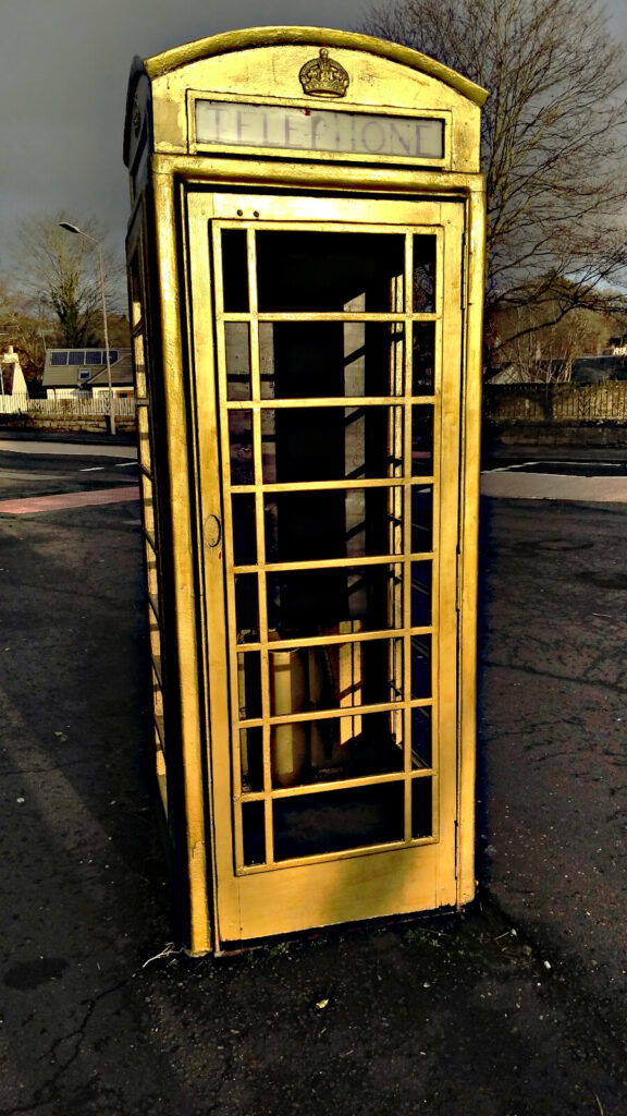 The golden telephone box in Dunblane is shown on a quiet street with very grey clouds in the background.
