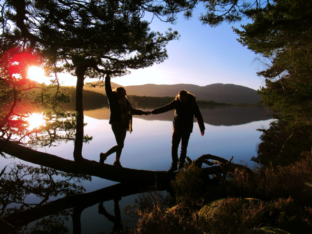 Zoe and boy-Zoe stood on a log on the edge of Loch Garten. Zoe is holding onto a tree branch with one hand and helping boy-Zoe up with the other. The sun has barely risen over the hills and a red glare hits the camera lenses. The water behind is perfectly still and a mirror reflection of the hills and sky.