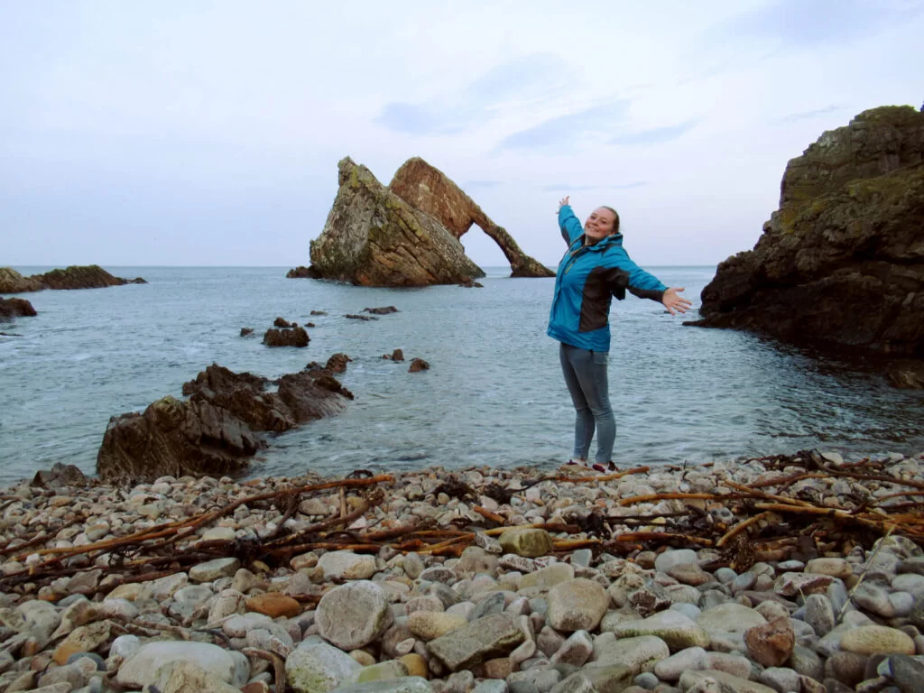 Bow Fiddle Rock is shown in the distance. Zoe is stood on the pebble beach, arms outstretched smiling at the camera.