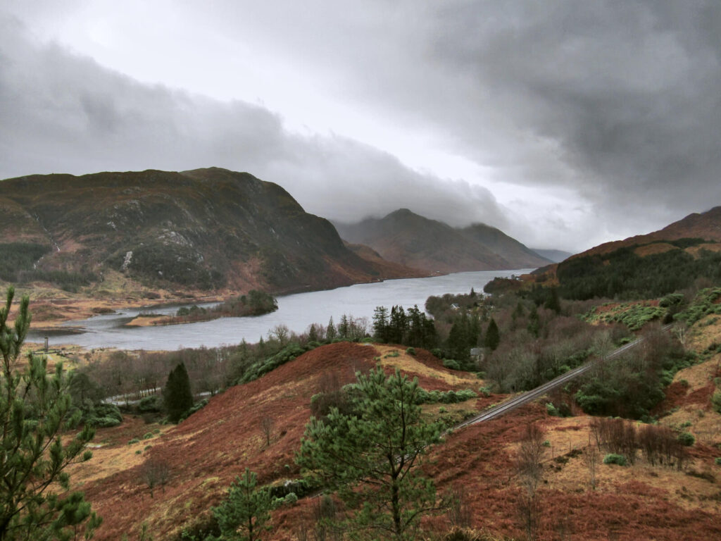 Clouds hang low over the hills and mountains surrounding Loch Shiel. The brown and red earth contrasts against the grey sky.