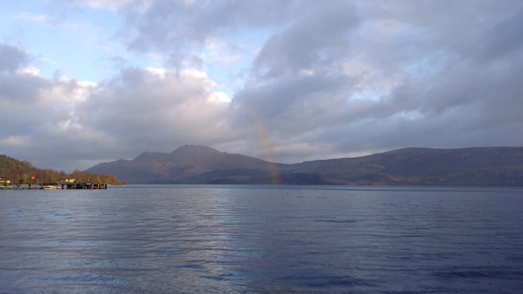 A rainbow emerges out of Loch Lomond and up into the clouds. It is a cloudy day but the sun is out. The hills on the opposite side of Loch Lomond can be seen.