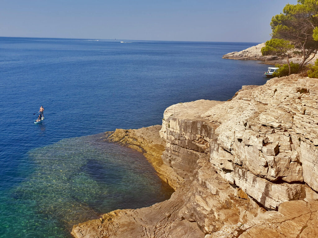 The photo is taken from the edge of a cliff looking down at the sea. The rocks are ragged as they enter the sea and are visible under the clear blue water