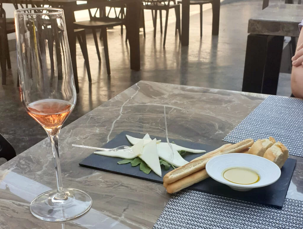 A small glass of rose wine is accompanied by cheese, breadsticks, sliced baguette and olive oil on a marble-look table. Pula is worth visiting for the wine it produces alone!