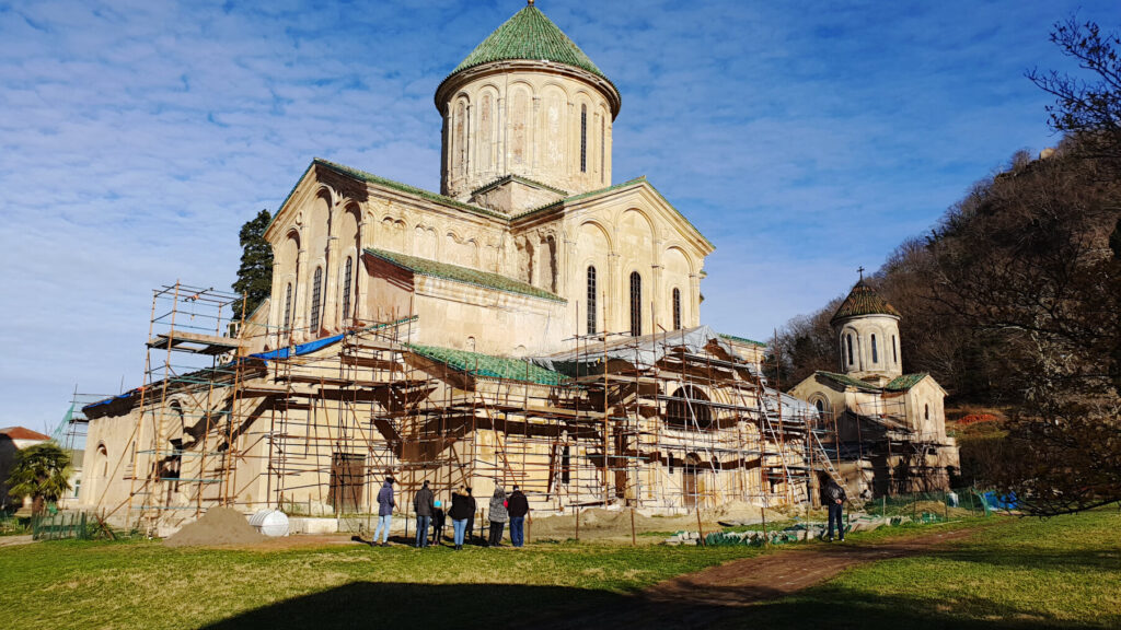 Gelati Monastery shown with scaffolding around the bottom of the building. A small group of visitors stand in front of the church admiring it's stunning medieval architecture.