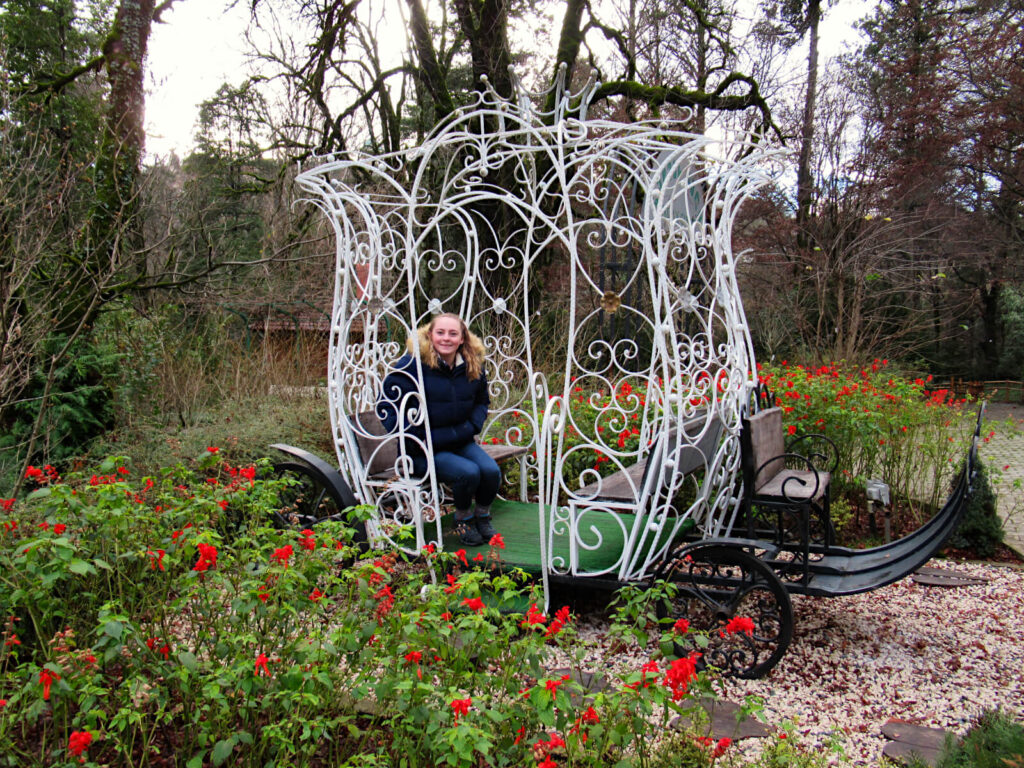 Zoe sits in a sculpture of a horse-drawn cart (no horses!), smiling at the camera. Although it is winter and many trees are bare, bright red flowers surround the carriage