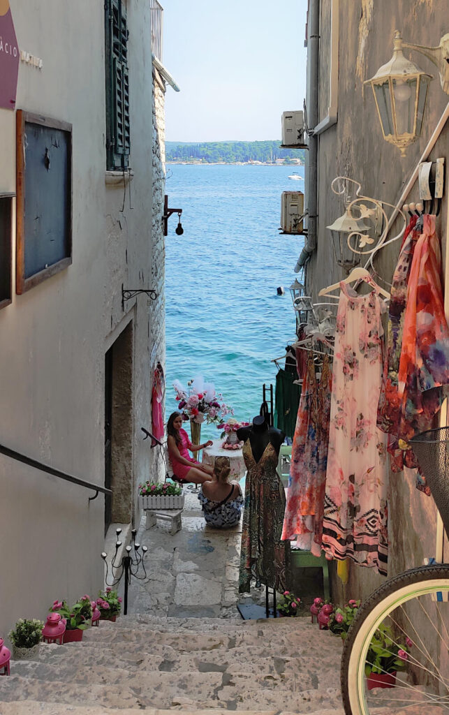 Steps are going down to a shop entrance, which sits between two tall buildings. The sea is directly behind the bottom of the stairs. At the centre of the photo two women are sat talking