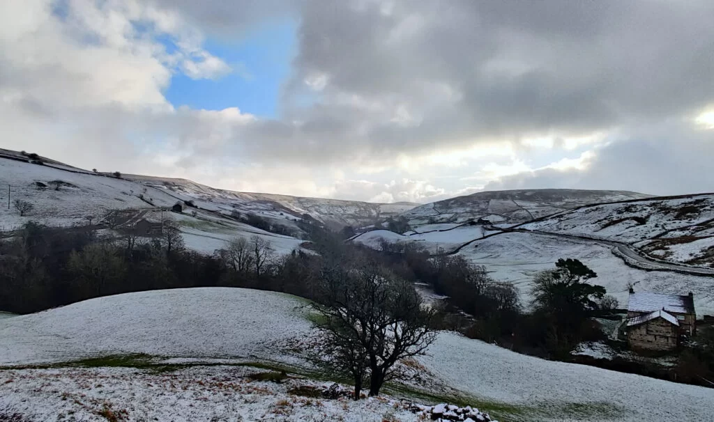 Looking back on this Yorkshire Dales Waterfall walk route. The track back to East Gill Force is on the left and the road back to Keld is on the right. The rolling hills of the Yorkshire Dales are shown under a light covering of snow.