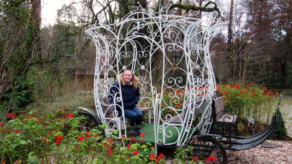 Zoe sits in a sculpture of a horse-drawn cart (no horses!), smiling at the camera. Although it is winter and many trees are bare, bright red flowers surround the carriage