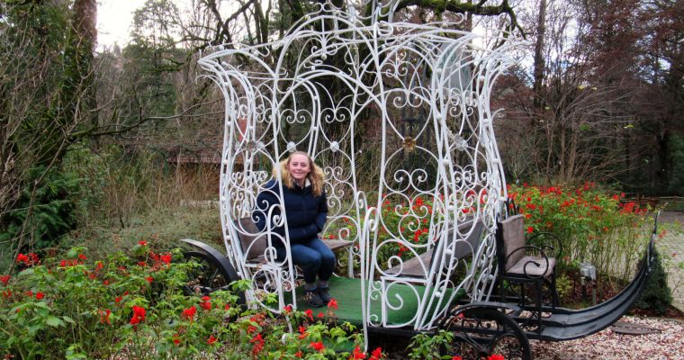 Zoe sits in a sculpture of a horse-drawn cart (no horses!), smiling at the camera. Although it is winter and many trees are bare, bright red flowers surround the carriage