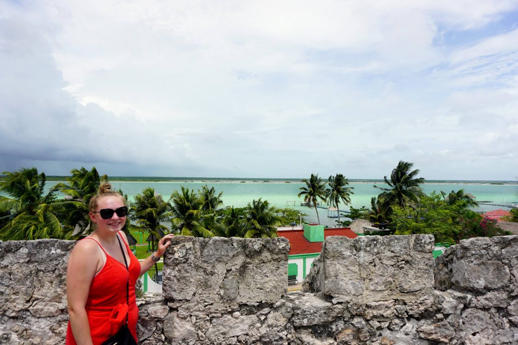 Zoe stood against the wall of Bacalar fort wearing a bright red jumpsuit. The bright colours of the lagoon are visible in the background