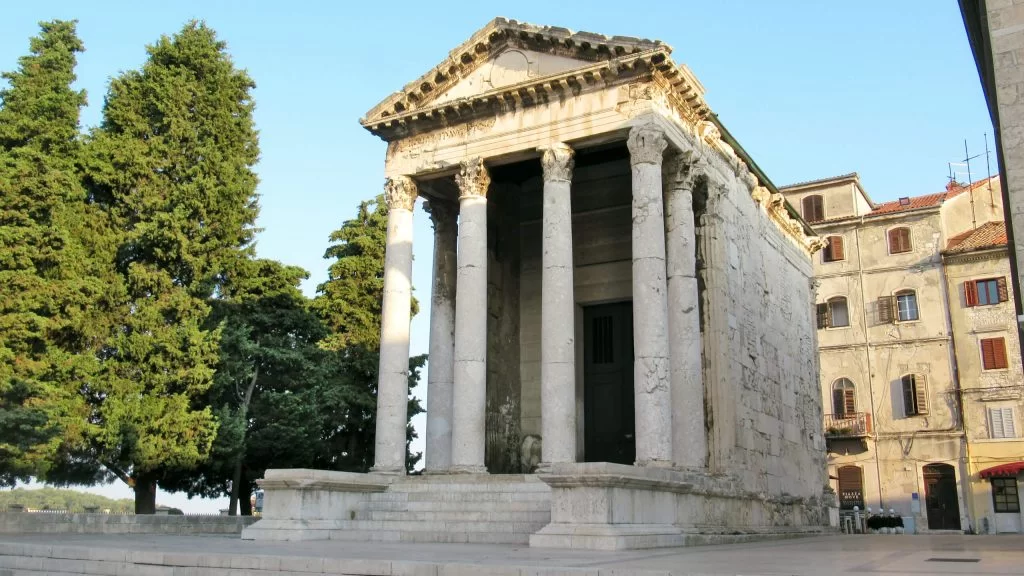 A roman-era temple with stairs leading up to the front four columns with a intricately patterned triangular roof. The building shows signs of it's 2000 year age but is otherwise well-preserved and not crumbling.