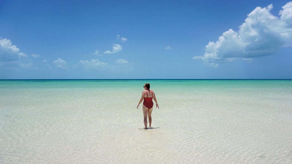 Punta Mosquito in Holbox is postcard paradise. In the image you can see a white sand beach covered by shallow clear water and a perfect blue sky. Zoe is stood in the centre of the image looking out to sea.