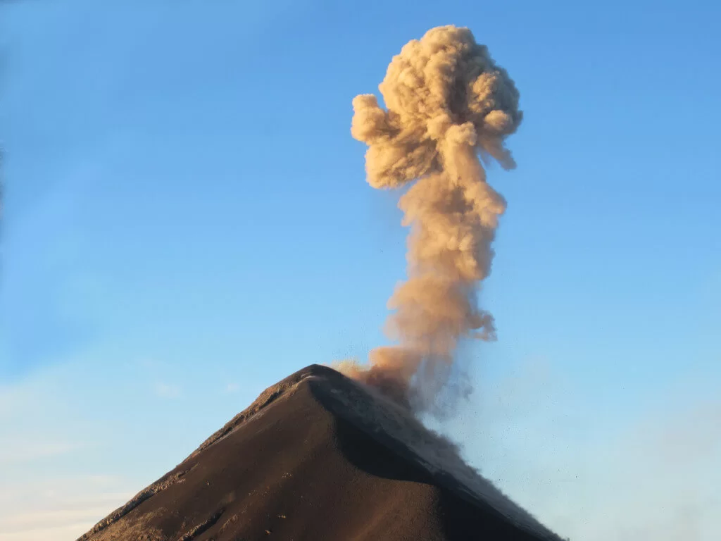 Volcano Fuego erupting during day light. A large blume of smoke is growing out of the volcano crater against the clear blue sky