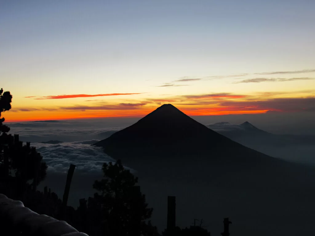 The silhouette of Volcan Agua as the sun starts to rise. The sky is orange and clouds cover the ground below.