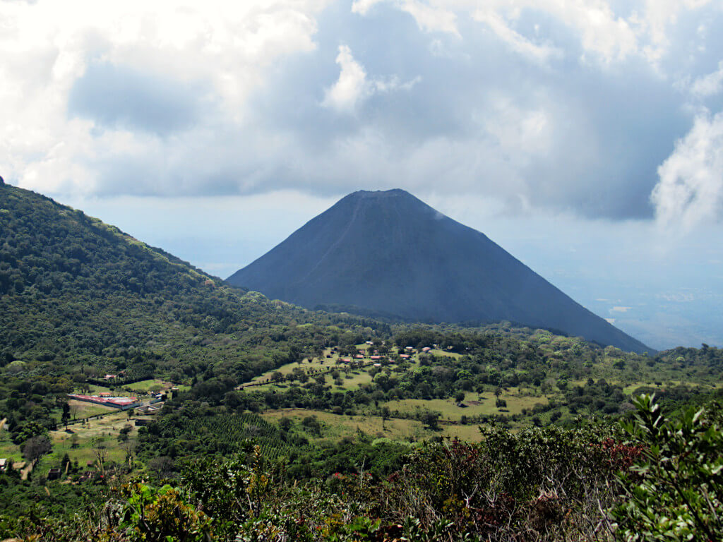 Looking towards Izalco volcano from the Santa Ana volcano trail. The volcano is dark and shadowed. In the foreground are lush green fields