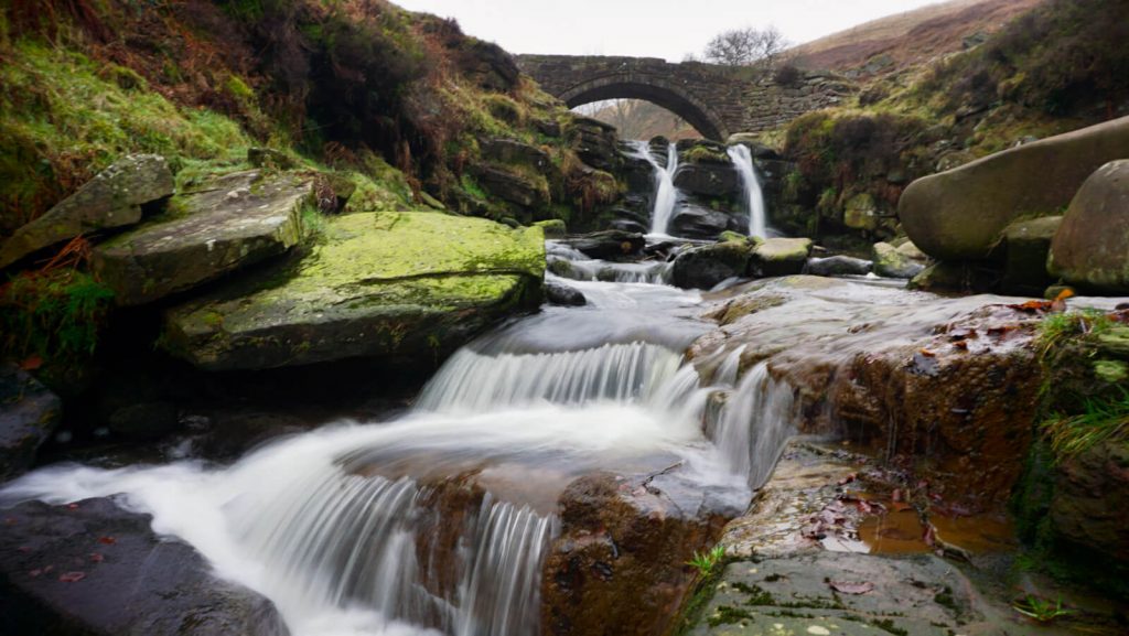Long exposure of water flowing over the mossy rocks at Three Shires Head with the bridge in the background