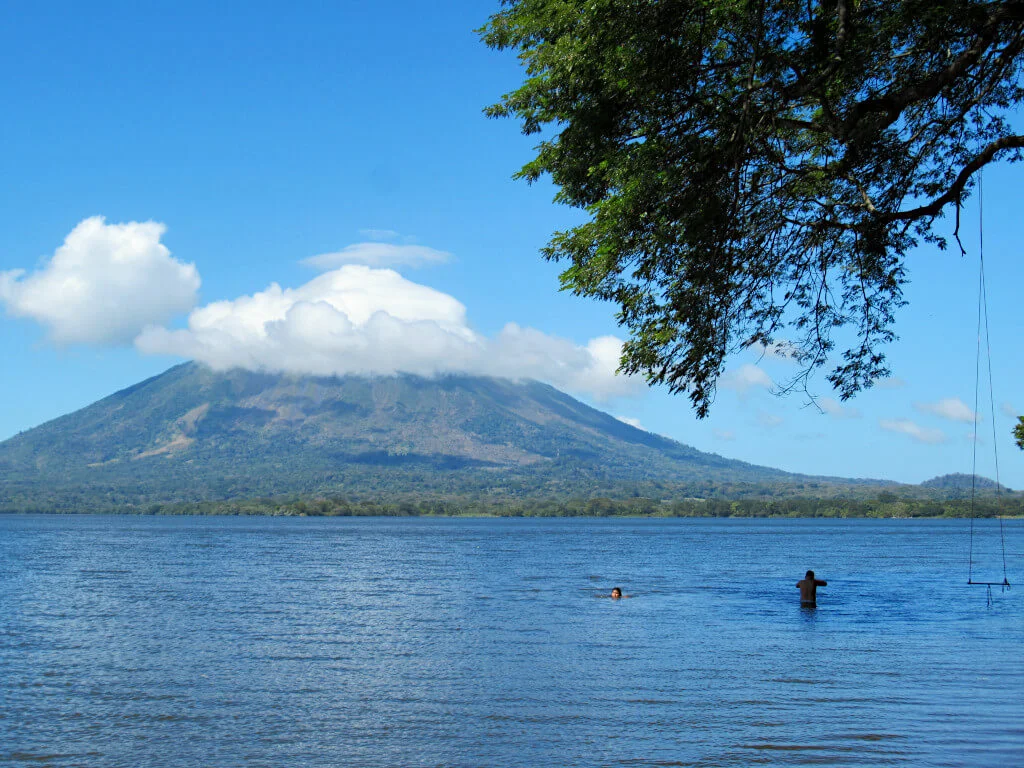The volcano partially covered by clouds in the background, the lake and two people swimming in it are in the foreground