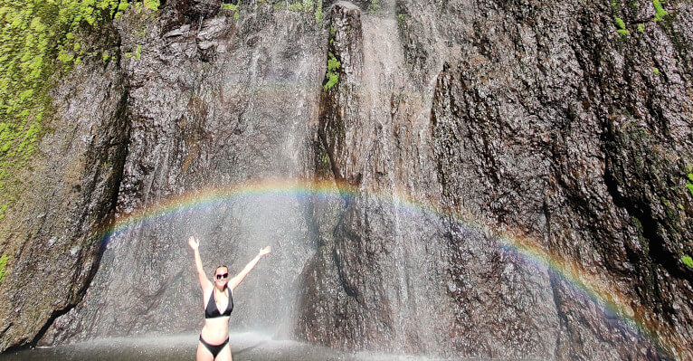 Zoe stood under a rainbow in the pool under the San Ramon waterfall on Ometepe. She is wearing a black bikini and has both arms in the air