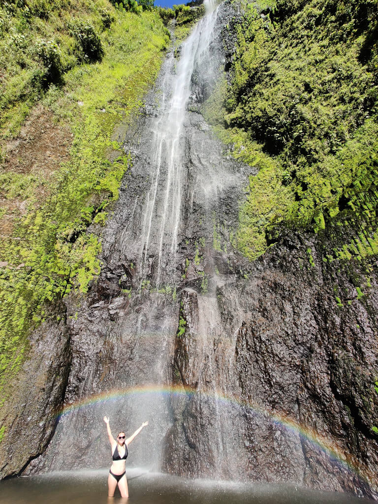 Zoe stood under a rainbow at the base of the waterfall, which towers above her. She is wearing a black bikini and has both arms in the air