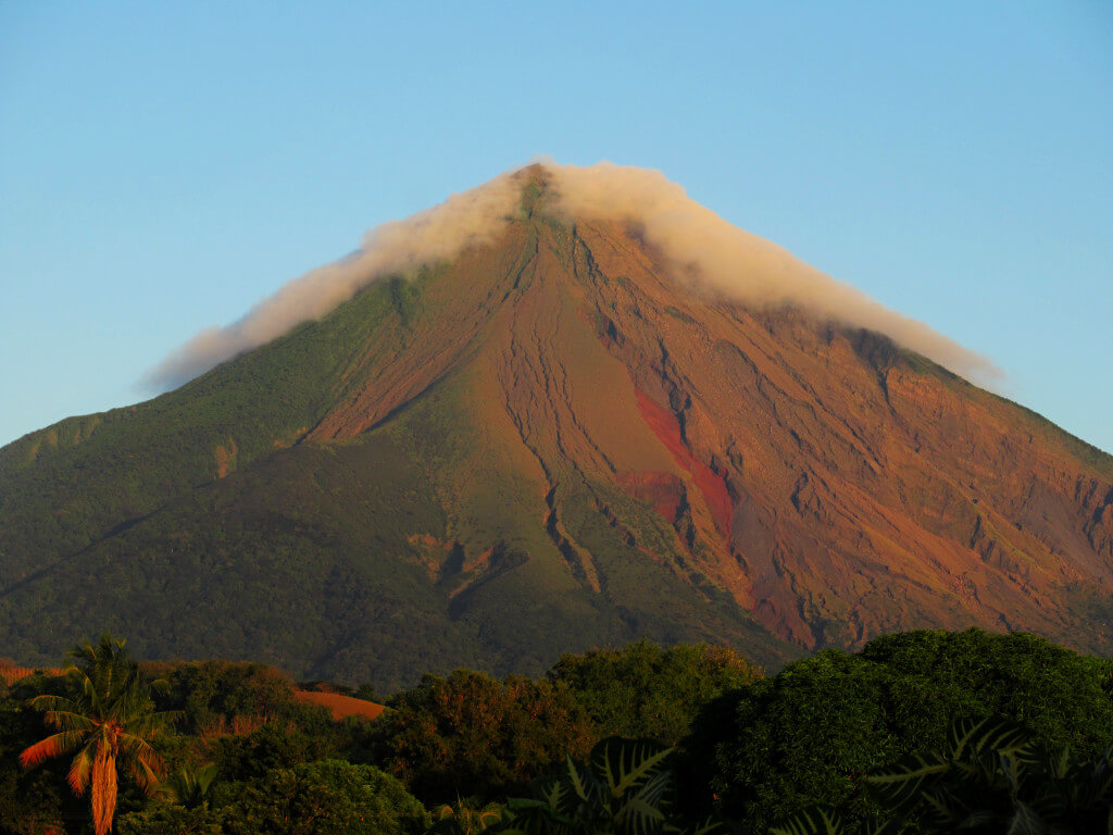 Volcano Concepcion on Ometepe's western side at golden hour. The volcano glows orange and green against the backdrop of a clear blue sky