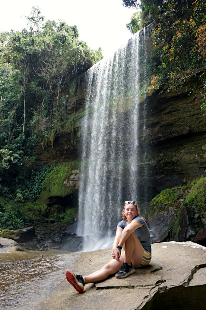 Zoe sits in front of Cascada El Perico on a large rock, smiling at the camera