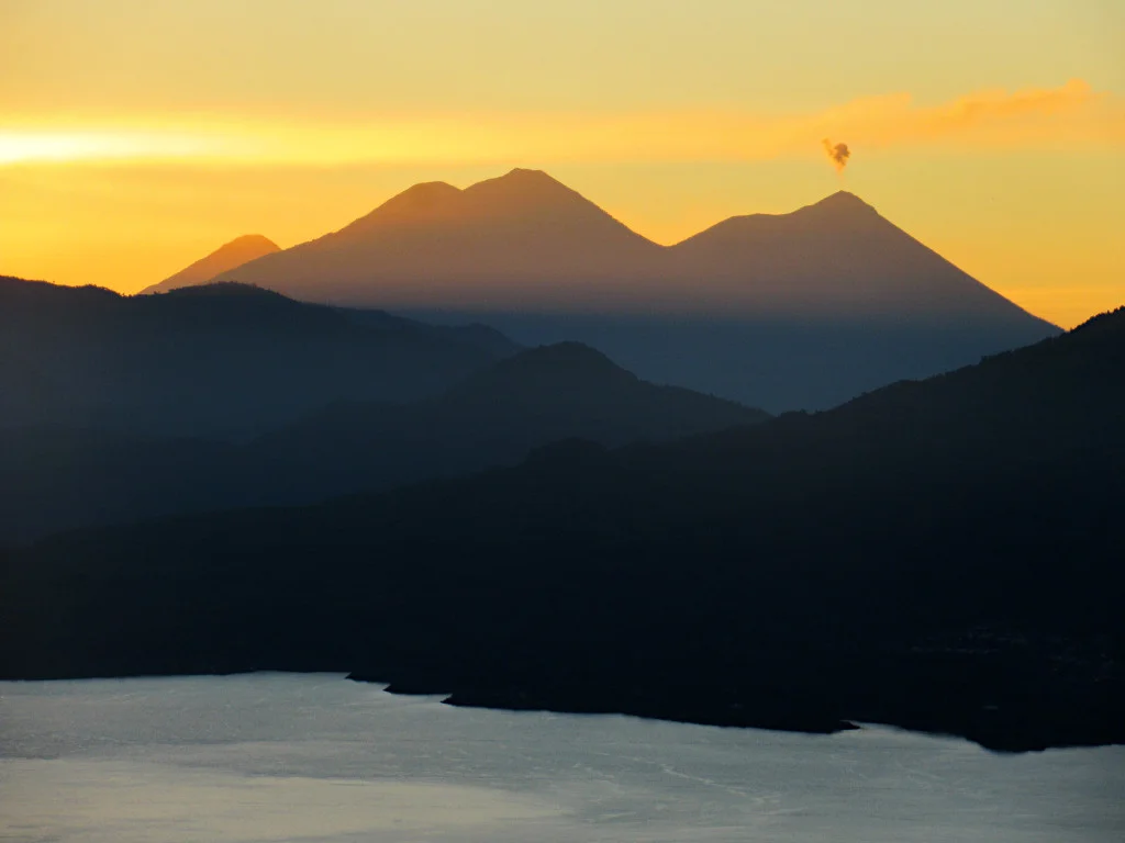 An ashcloud spews from Fuego against a yellow sunrise sky with Lake Atitlan in the foreground. Acatenango and Agua are also visible in the early morning sky