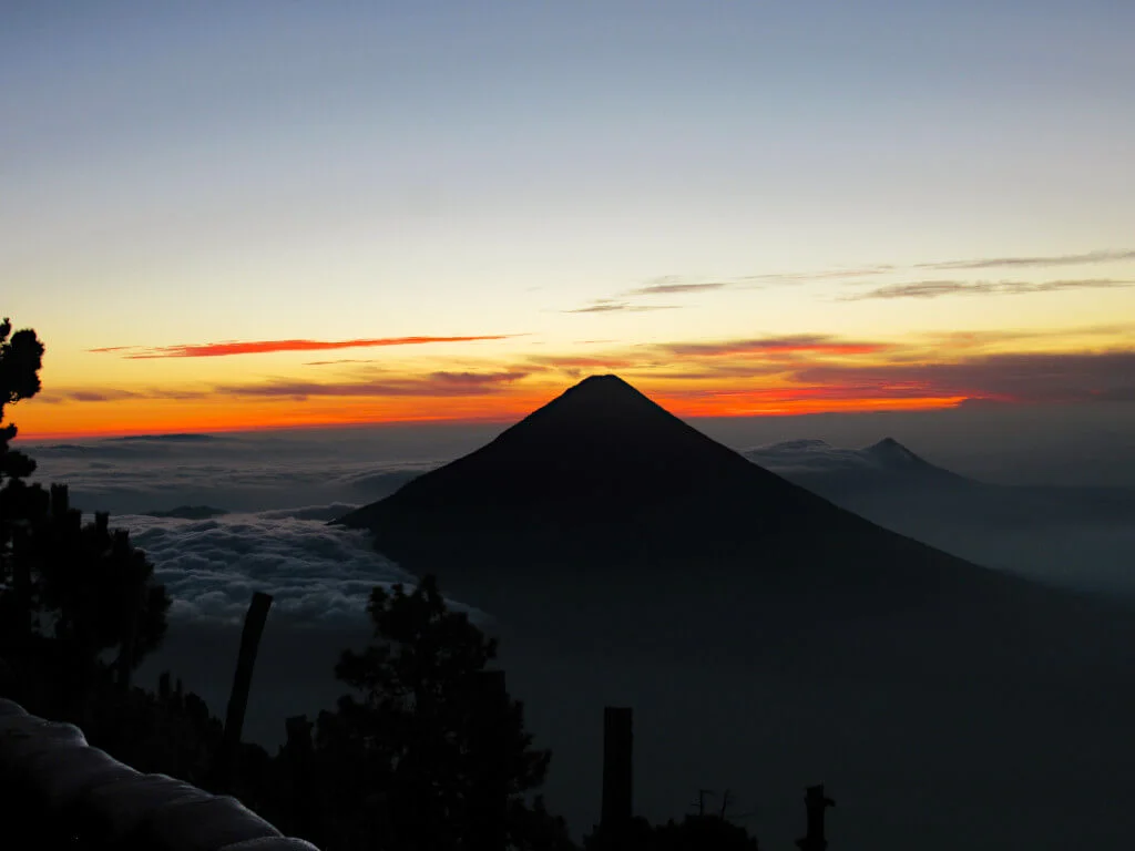 Volcano Agua and Pacaya in the distance as silhouettes as the sun rises behind them, photographed from the base camp area of Acatenango