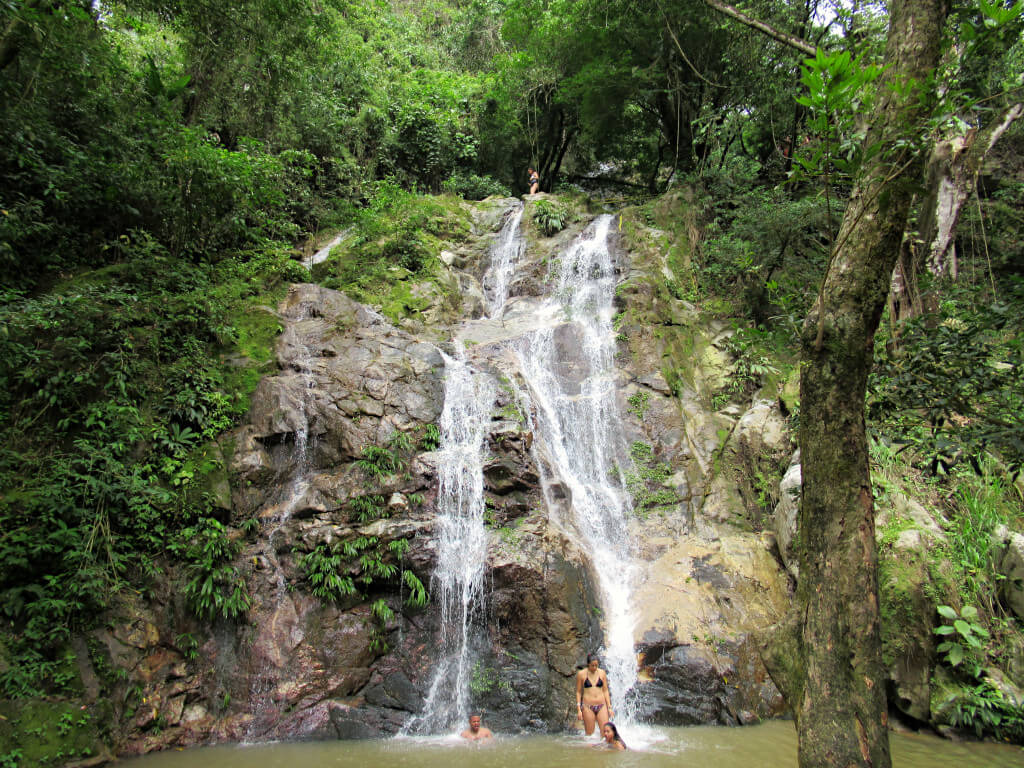 Three people cool off in the pool in front of Marinka Waterfalls in Minca on the northern coast of Colombia. The water runs close to vertical down across rocks from height