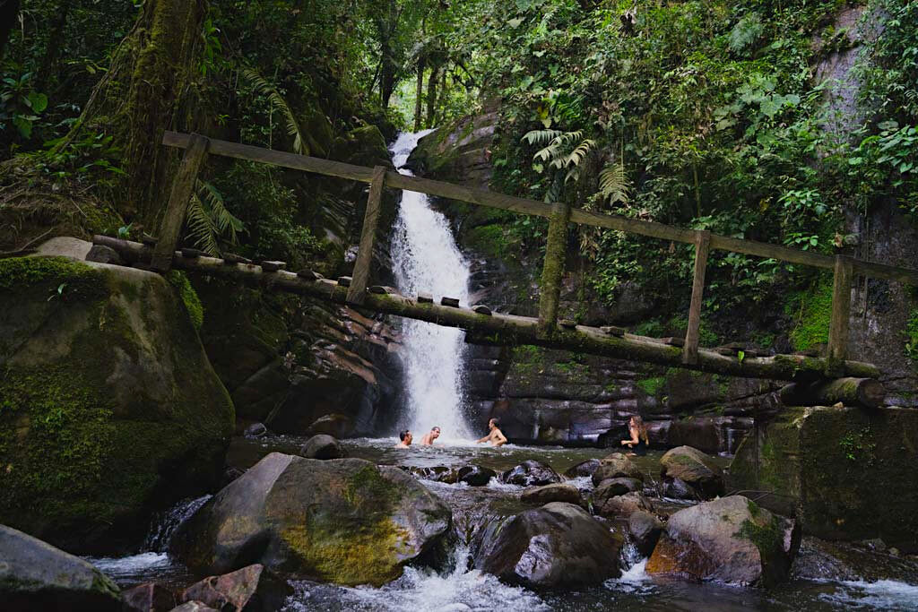 A wooden bring lies between rocks in front of the Santa Rita Waterfall, not far from Salento. 4 people are swimming in the pool below the waterfall surrounded by greenery