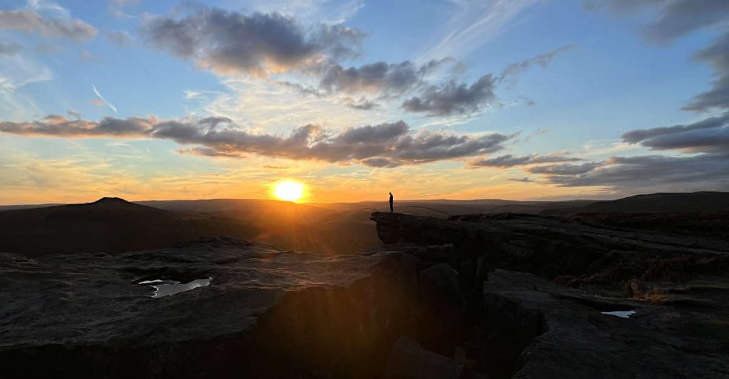 A single figure stands on the rocky overhang of Bamford Edge walk as the sun sinks beneath the hills behind