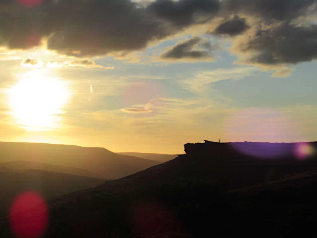 A lone walker stands on Bamford Edge as the sun sets over the Derbyshire hills
