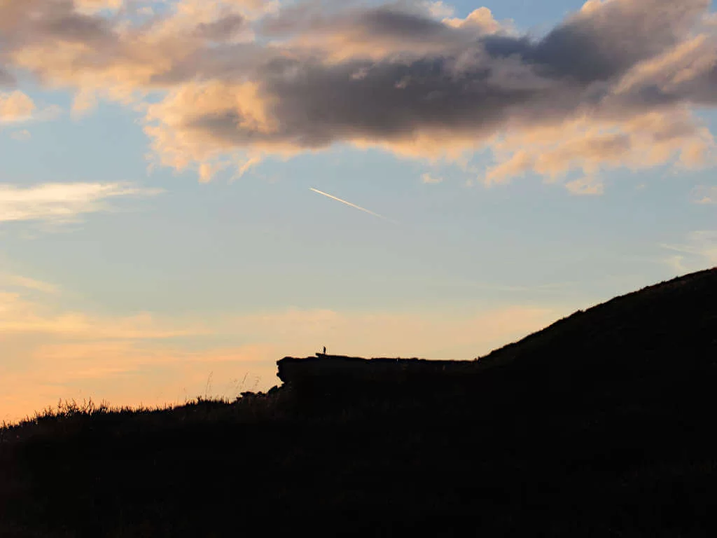 A lone walker stands on the edge of Bamford Edge in silhouette against a sunset sky with a plane flying high overhead