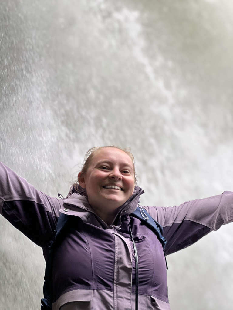 Zoe smiling at the camera with both arms in the air, stood in front of a waterfall that takes up the whole frame
