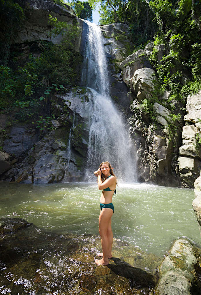 A young woman wearing a green bikini stands in front of Yelapa Waterfall, the water is a light shade of green and rocks surround the waterfall pool.