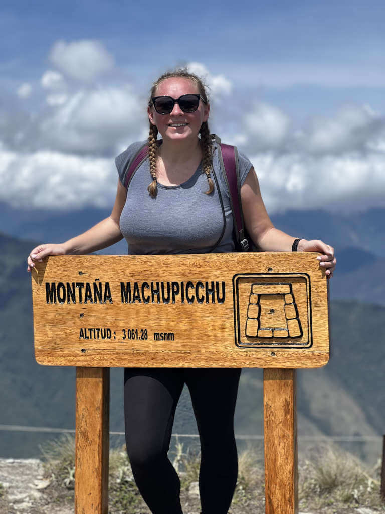 Zoe stood behind a sign, smiling, that reads 'Montana Machupicchu, altitude 3061.28 metres above sea level'