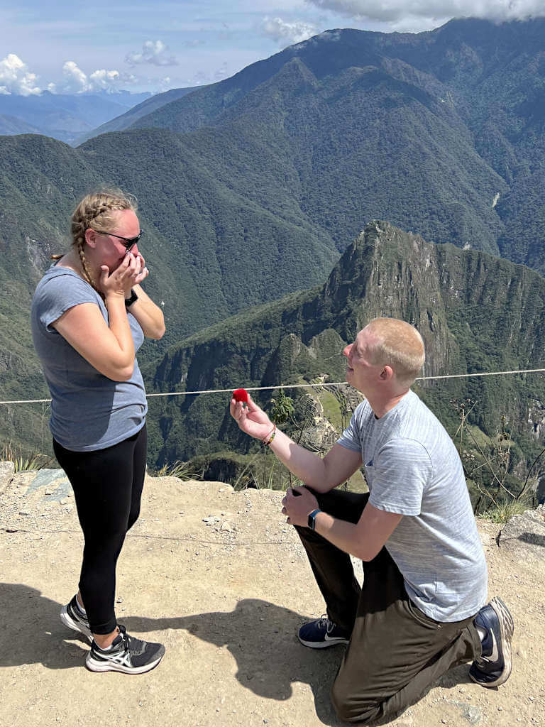 A man on one knee proposing to his girlfriend (me) who has both hands on her face in shock. The World Wonder ruins are in the background below