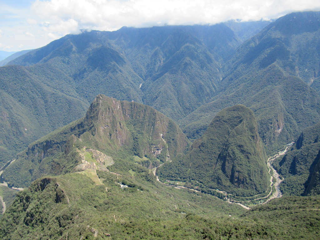 The ruins of Machu Picchu on the left and the river leading to Aguas Calientes on the right, the rest of the image is filled with large, tree-covered mountains