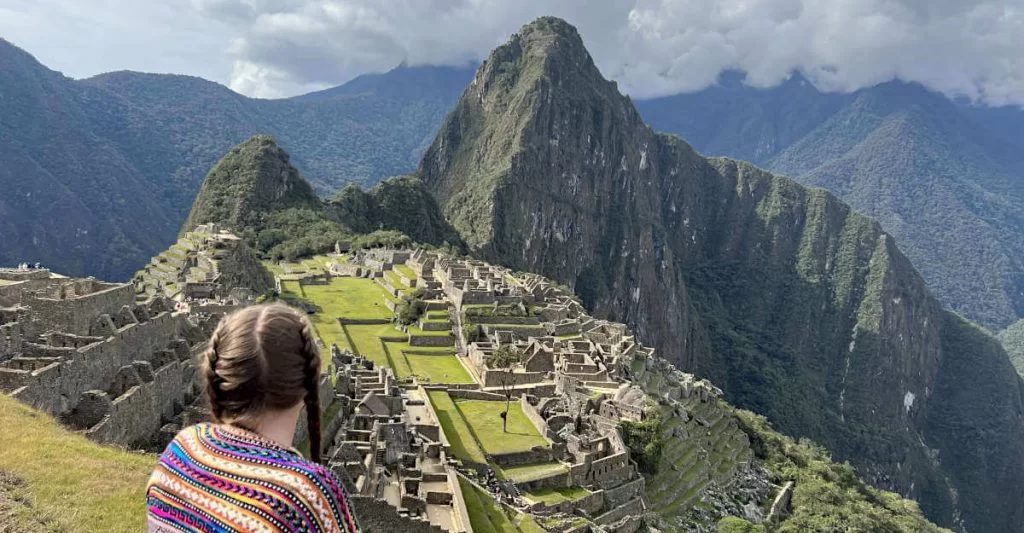 Zoe stood looking out over the ruins of Machu Picchu on the mountain top. She is wearing a pink Peruvian jumper with a colourful pattern and has her hair in two plaits.