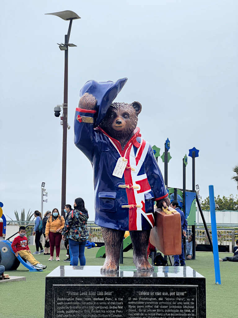 Paddington Bear statue in Lima. Paddington is wearing a Union Jack raincoat and a blue hat, carrying a brown suitcase