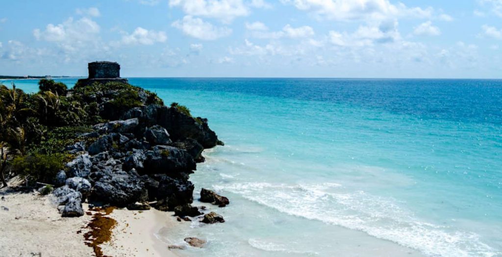 The vivid blues of the Caribbean Sea with Tulum ruins to one side. The gold sand beach is also visible in the bottom-left corner