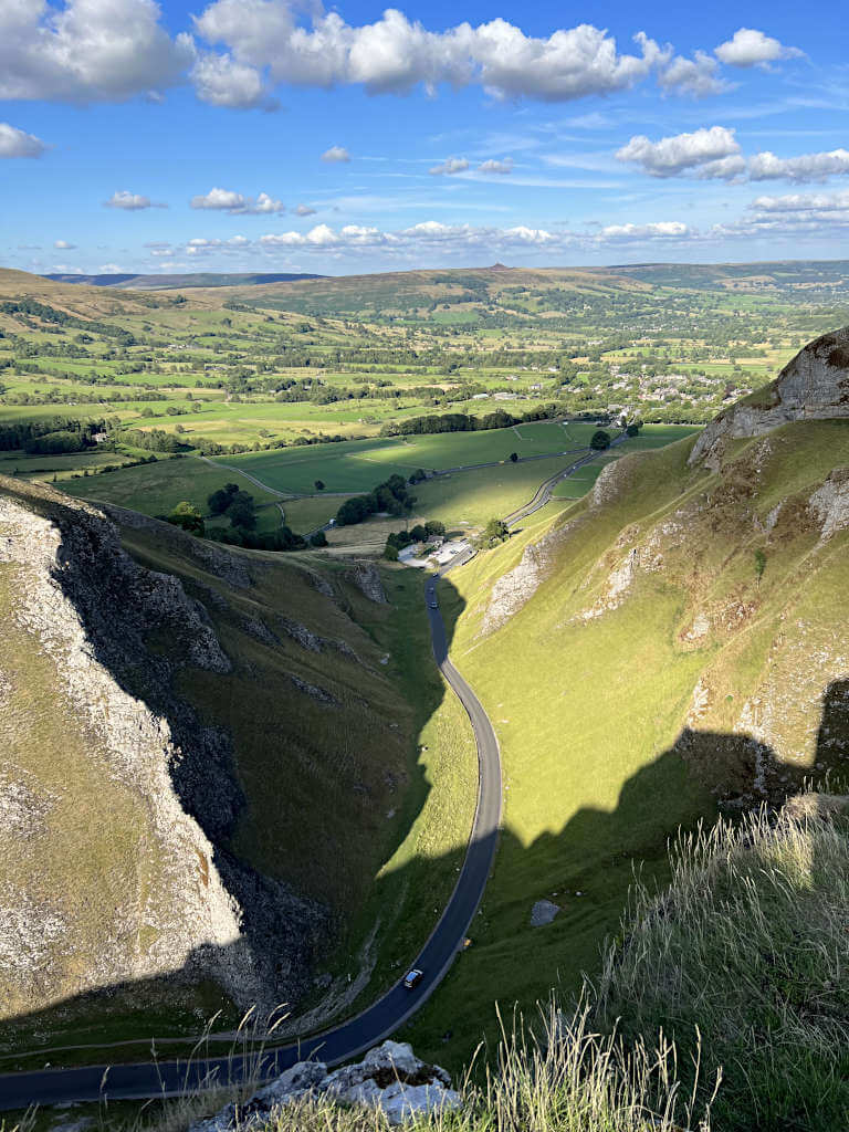 Looking along Winnats Pass from the rocky top towards Castleton with near endless green fields in the background under a blue sky with fluffy white clouds