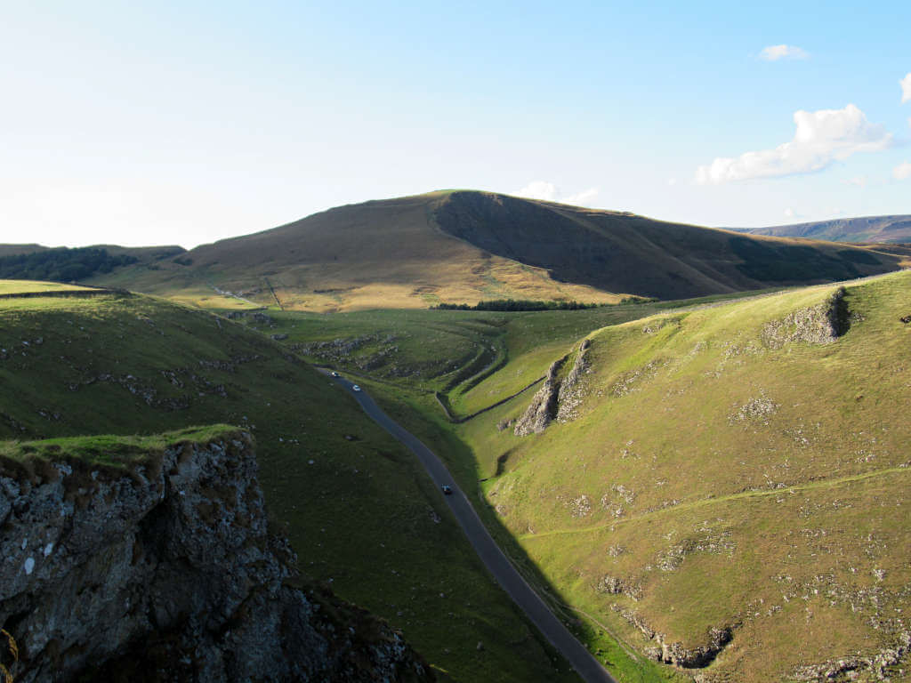Mam Tor towers over Winnats Pass as cars that appear tiny drive through the steep valley below