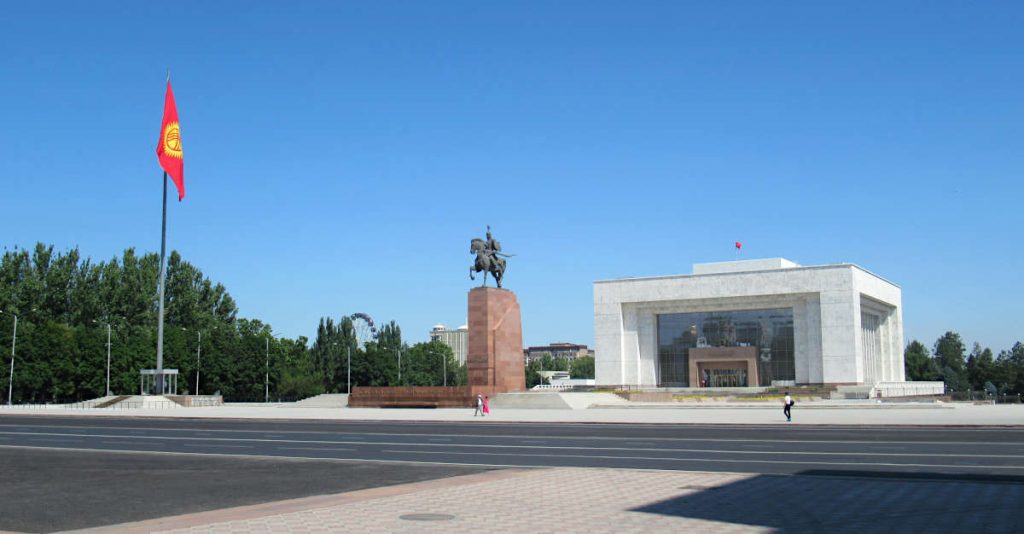 Ala-Too Square Bishkek with a perfect blue sky and just a couple of passers by. The flag waves gently in the wind with the statue and large building also visible