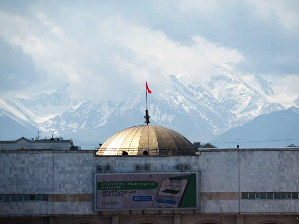 The snow-covered mountains in the distance behind a building with a Kyrgyzstan flag on top in the city,