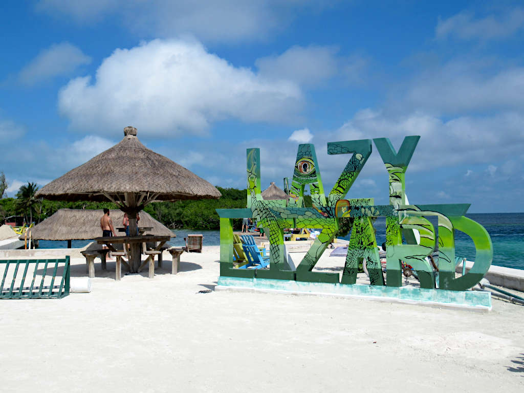 Large letters that spell out Lazy Lizard that have a lizard design painted on them, next to a palapa and the sea