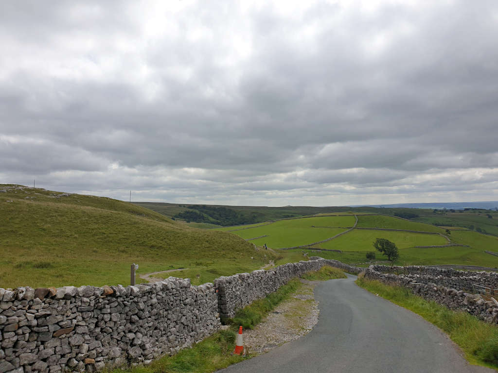 A single-track country road with a drystone wall on either side and rolling Yorkshire hills in the background