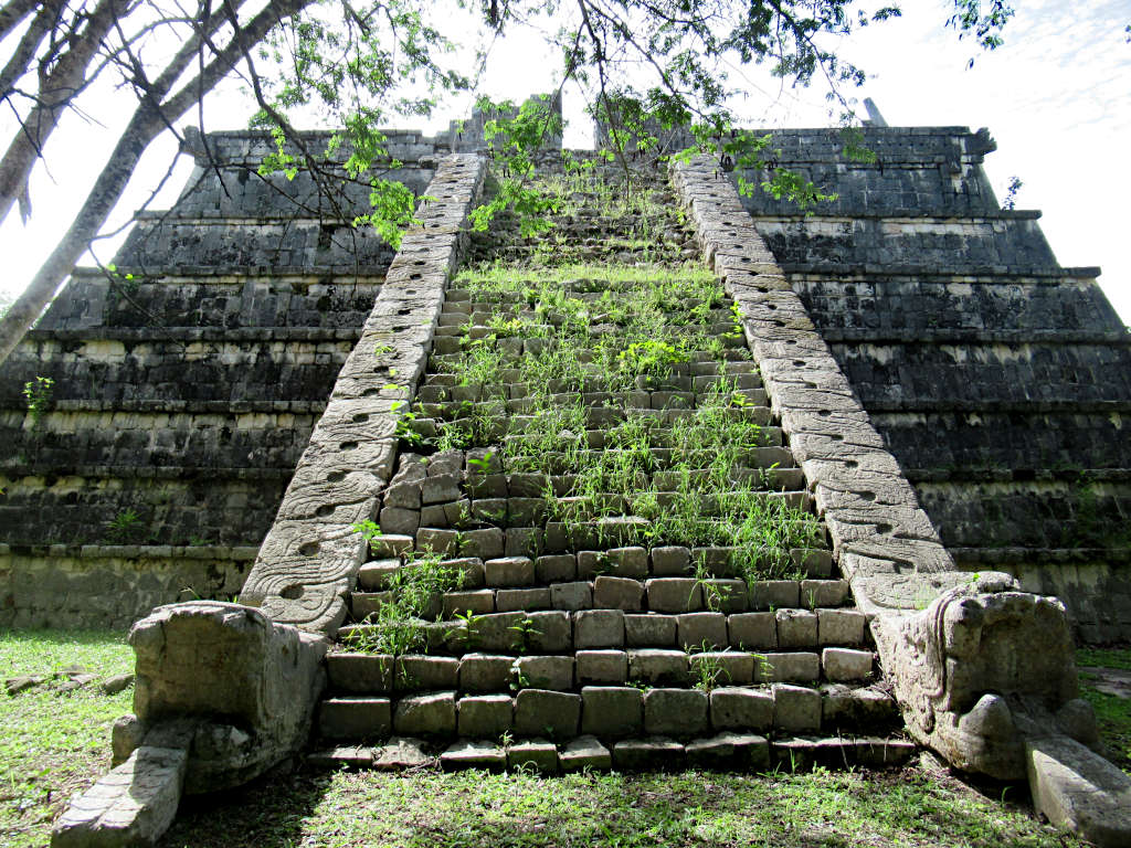 A small pyramid temple at Chichen Itza. Weeds grow through the steps that lead up to the top and a tree hangs over the top of the frame